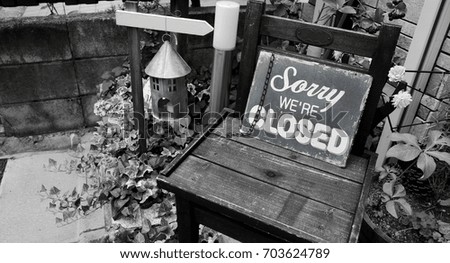 Closed Sign (Sorry we are Closed) Blue Metal Board Lean Against on Wooden Chair among Pot of Plant in Black and White 