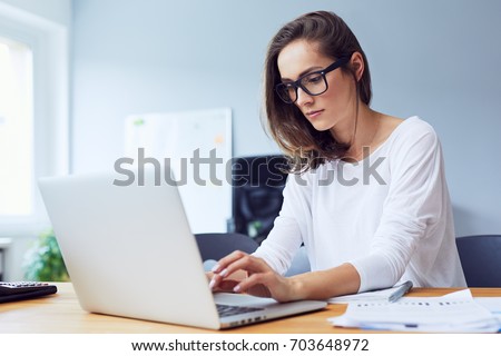 Concentrated young beautiful businesswoman working on laptop in bright modern office Royalty-Free Stock Photo #703648972