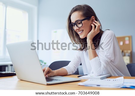Portrait of beautiful cheerful young businesswoman working on laptop and laughing in home office Royalty-Free Stock Photo #703649008