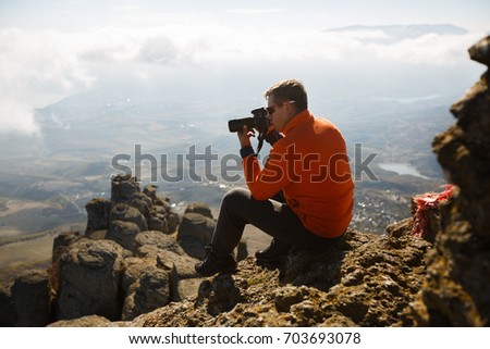 Young professional traveller man with dslr camera shooting outdoor fantastic mountain landscape. Hiker sits on a rock at the high summit and photographing above clouds