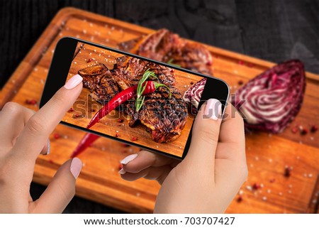 Photographing food concept - tourist takes picture of ready-to-eat beef steak dish on cutting wooden board on smartphone.