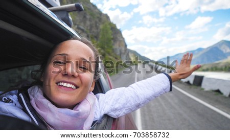 Smiling young brunette woman in a car playing with wind and driving past the beautiful mountains