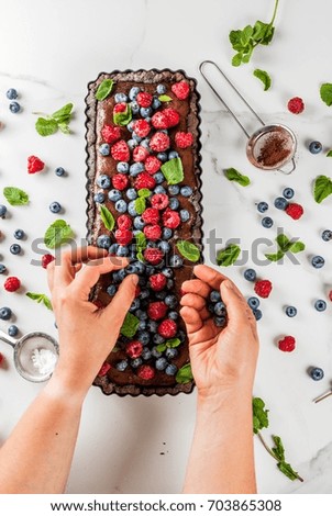 Summer homemade baked pastry. Chocolate cake tart with chocolate cream, woman decorate it with blueberry raspberry mint leaves, powdered sugar. White marble table, copy space top view hands in picture
