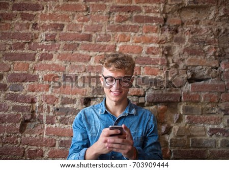 Stylish happy hipster guy chatting with friends in social network via cell telephone, sitting against brick wall. Young smiling man student typing text in internet via modern mobile phone