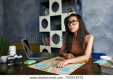 Beautiful serious young Asian female wearing glasses sitting at her workplace, pointing finger on world map, choosing destination while having appointment with client. Tourism, traveling and planning