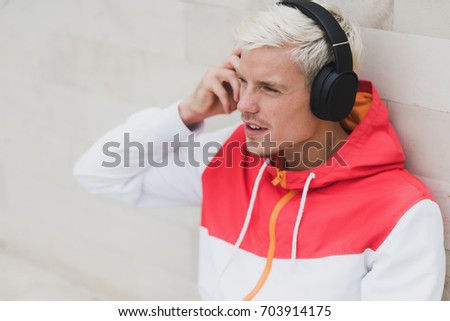 Enjoying attractive portrait of young male dressed in red sportswear resting after physical activities in open air in park.Young handsome man listening to music with headphones against grey background