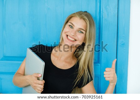 Portrait of a young beautiful girl with blond hair with a laptop on a blue background. The girl is showing her thumb up. Joy, fun and happiness concept. Portrait of happiness.Positive human emotions 
