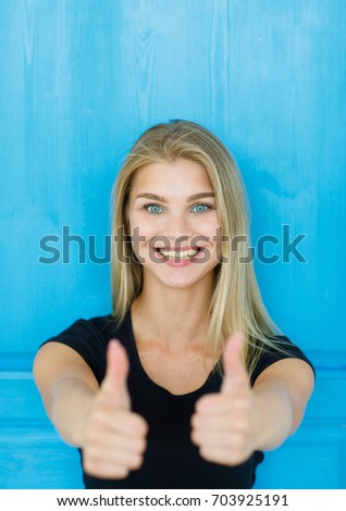 Happy young woman giving thumbs up on blue background.Cheerful beautiful girl in black T-shirt showing thumbs up.Portrait of a young woman showing thumbs up as a sign of success.Portrait of happiness
