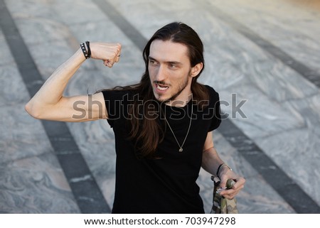 Outdoor lifestyle picture of funny young Caucasian man with beard and long dark hair having fun on city streets, looking sideways, showing off his bicep in front of friends. Youth and urban life