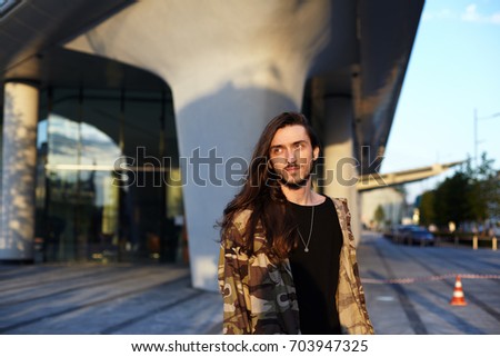 Urban fashion portrait of attractive trendy-looking young bearded Caucasian man dressed in camouflage bomber over black t-shirt, having nice walk on city streets with friends on sunny warm evening