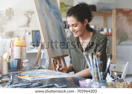 Picture of cute female artist sitting at table, surrounded with watercolors, drawing something at easel, having happy expression. Brunette young woman being busy with creative work at workshop