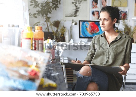 Portrait of thoughtful beautiful young brunette female artist wearing khaki shirt and ripped jeans sitting on chair at home workshop, looking through the window, having inspired expression on her face