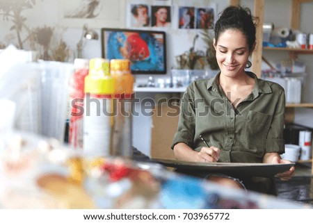 Good-looking cheerful professional young female artist working on new creative project, drawing, making sketches with pencil, feeling inspired. People, job, occupation, profession and hobby concept