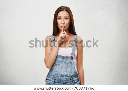 Secrecy, conspiracy gossiping and confident information. Picture of beautiful brunette girl in stylish jeans jumpsuit, holding finger on lips as silent gesture, asking to keep silent about her secret