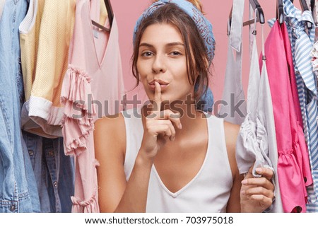 Adorable woman in casual clothes, showing silence sign while standing near wardrobe with clothes, demonstrating conspiracy. Woman choosing trendy clothes on ranks. Beauty and fashion concept