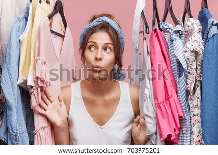 Attractive woman dressed casually, looking with doubts aside while standing near hangers with clothes, thinking what to dress on business meeting with companions. Woman of fashion having many clothes Royalty-Free Stock Photo #703975201