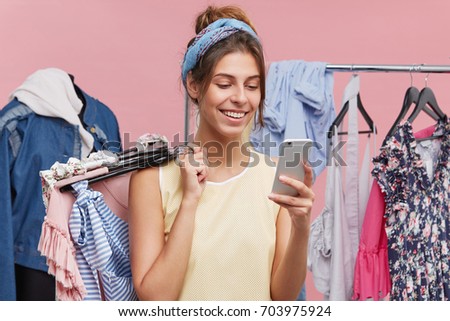 Happy female standing at clothing store, messaging with friend over smart phone while trying new clothes asking for advice what to buy. Cheerful woman using modern cell phone in shopping mall.