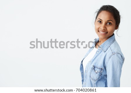 People and beauty concept. Portrait of beautiful dark-skinned woman posing on white background looking at camera with cute and charming smile, dressed in light-blue denim shirt.