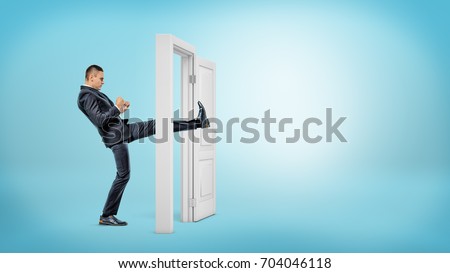 A businessman in side view kicks a small white door open with his leg on blue backgrounds. Business and success. Opening all doors. Aggressive business approach. Royalty-Free Stock Photo #704046118