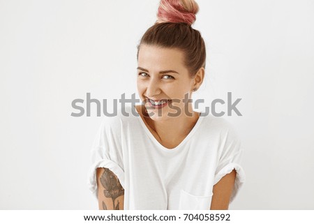 Close up indoor shot of attractive charming young tattooed Caucasian female with mysterious blue eyes wearing pink hair tied in knot, posing at studio wall, dressed in white oversize t-shirt
