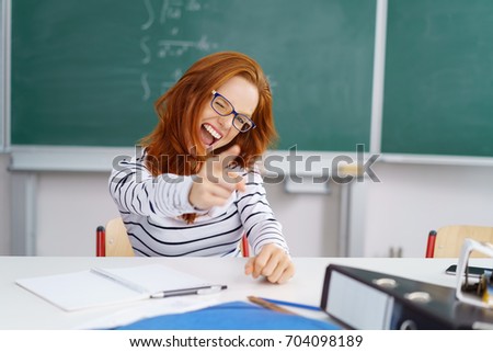 Vivacious young student giving a thumbs up of success and cheering as she sits at a desk in the classroom working