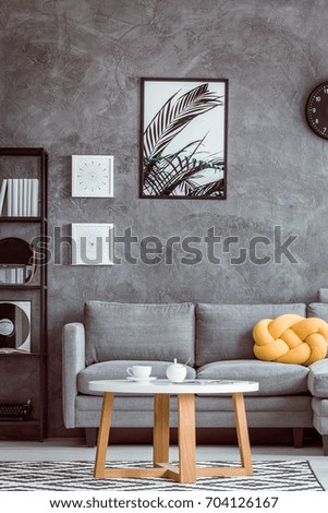 Painting of leaves on dark wall above grey settee with yellow pillow in simple living room