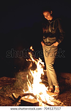 Young woman in geans and hoodie in the mountains. Young woman with fire on mountain at sunset. Girl standing near a fireplace