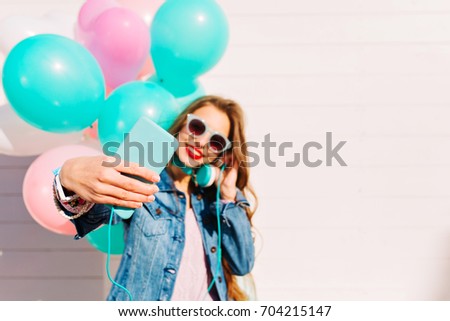 Adorable young long-haired woman with balloons makes selfie on smartphone while listening music. Portrait of happy brunette girl enjoing favourites song in headphones and taking picture of herself
