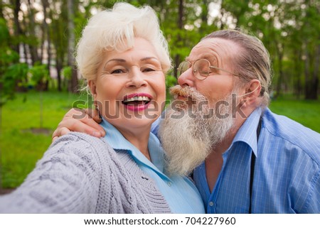 An elderly couple in love kisses in a green park with flowers and making selfie
