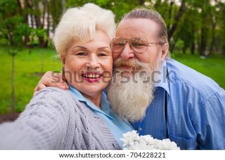 An elderly couple in love hugs in a green park with flowers and making selfie