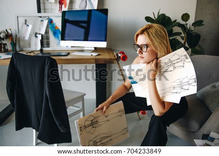 young pensive fashion designer holding sketches and looking away in office