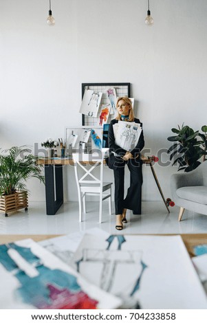 young fashion designer holding sketches while sitting on table and looking away in office