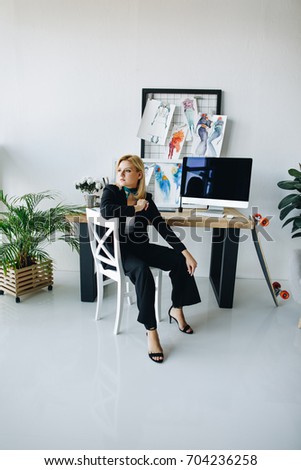 beautiful young fashion designer looking away while sitting on stool at workplace 
