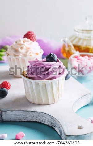 Colorful cupcakes with butter-cream and fresh berries on cutting board. Tea pot with green tea and pink rose sugar on background. Closeup view, toned image