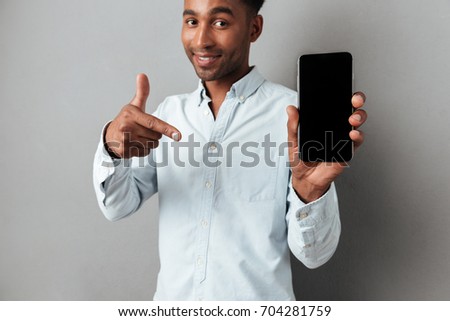 Excited cheerful african man pointing finger at blank screen mobile phone isolated over gray background