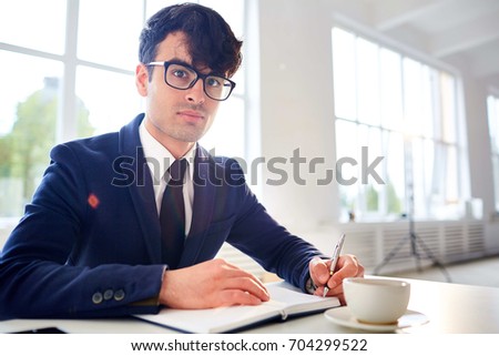 Handsome young businessman posing for photography while sitting at desk and taking notes, interior of spacious office with panoramic windows on background