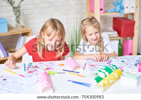 Portrait of two laughing little girls enjoying art class siting at desk and coloring pictures together