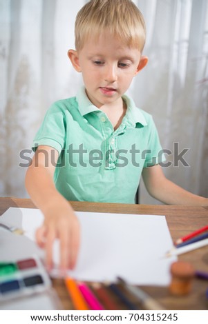 A little blond boy with blue eyes draws colored pencils on paper, a blank sheet on the table