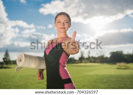 selective focus of smiling woman with yoga mat showing thumb up while standing on green lawn 