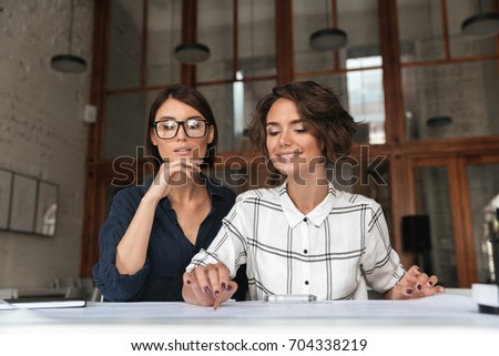 Two beauty pretty smiling women sitting by the table in co working office over glass doors background
