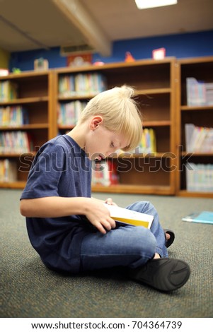 A little second grade elementary school aged boy child is sitting on the carpet, reading a book at the school library. Royalty-Free Stock Photo #704364739