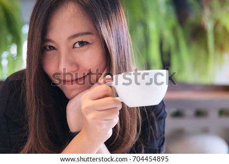 Closeup image of a beautiful Asian woman holding a white mug and drinking hot coffee with feeling happy in modern cafe and green nature background