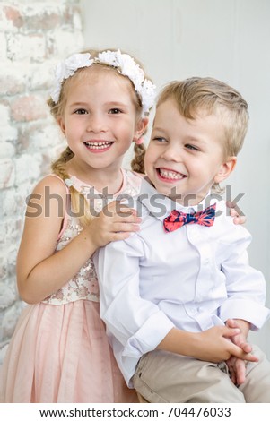 Two charming children stand having joined hands. Girl is dressed in a elegant dress. On the head of her a wreath. Boy has a bowtie. Children laugh.