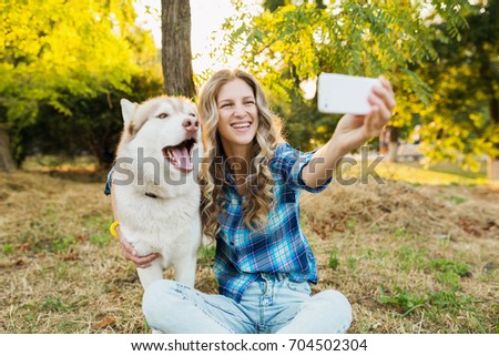 young beautiful woman playing with dog in park, summer lifestyle, casual style, sunny, smiling, happy, positive, husky breed, making selfie photo on smartphone, backlight