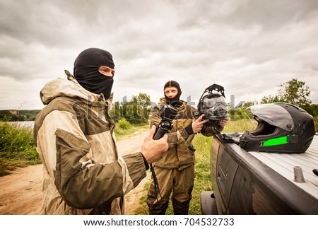 Two men outdoors wearing balaclava helmets and motorcycle uniforms, one man is holding sports action camera and another man is holding a helmet.