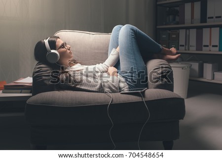 Young woman with headphones relaxing at home late at night, she is lying on the armchair and listening to music using a tablet Royalty-Free Stock Photo #704548654