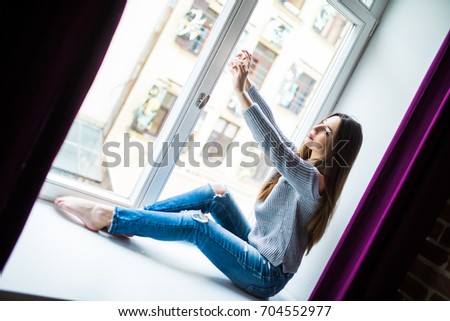 beautiful girl in casual clothes doing selfie using a smartphone and smiling while sitting on the window-sill at home