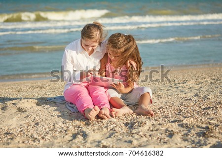 Cute Girl seventeen-year-old with Down syndrome and little girl on the beach play with the tablet. Positive human emotions, joy.