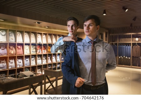 One elegant man helps another try on a suit in a clothing store. In the background a large regiment with shirts.