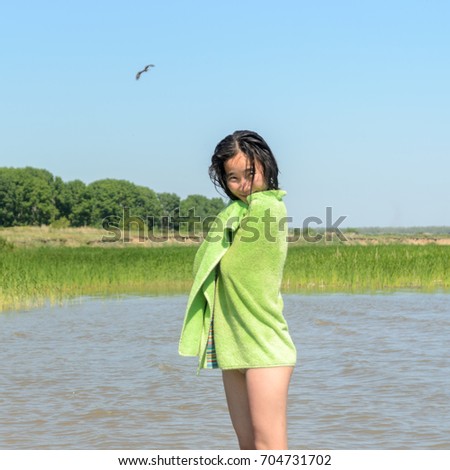 Beautiful Asian girl smiling is wrapped in green towel with wet hair in swimsuit on the background of the birds the eagle and the trees of the forest to the cliff and grass near the water. Soft Focus